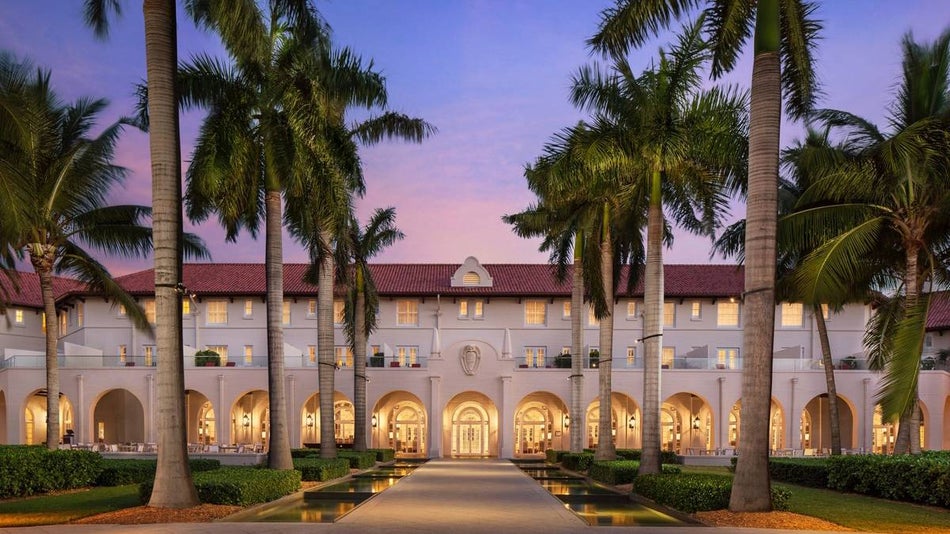 Wide shot of the Casa Marina, A Waldorf Astoria Resort with a walkway lined with palm trees and a pink purple sunset behind it in Miami, Florida, USA