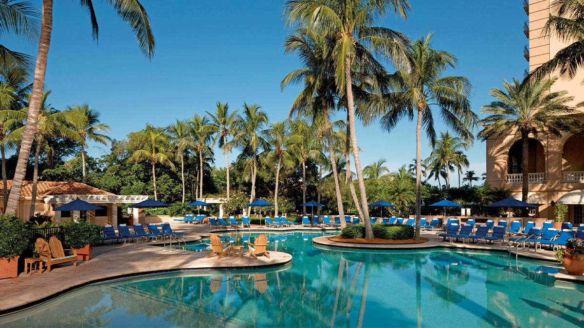 A bright blue pool lined with chairs for laying out on and palm trees for shade on a sunny day at The Ritz-Carlton Naples in Miami, Florida, USA