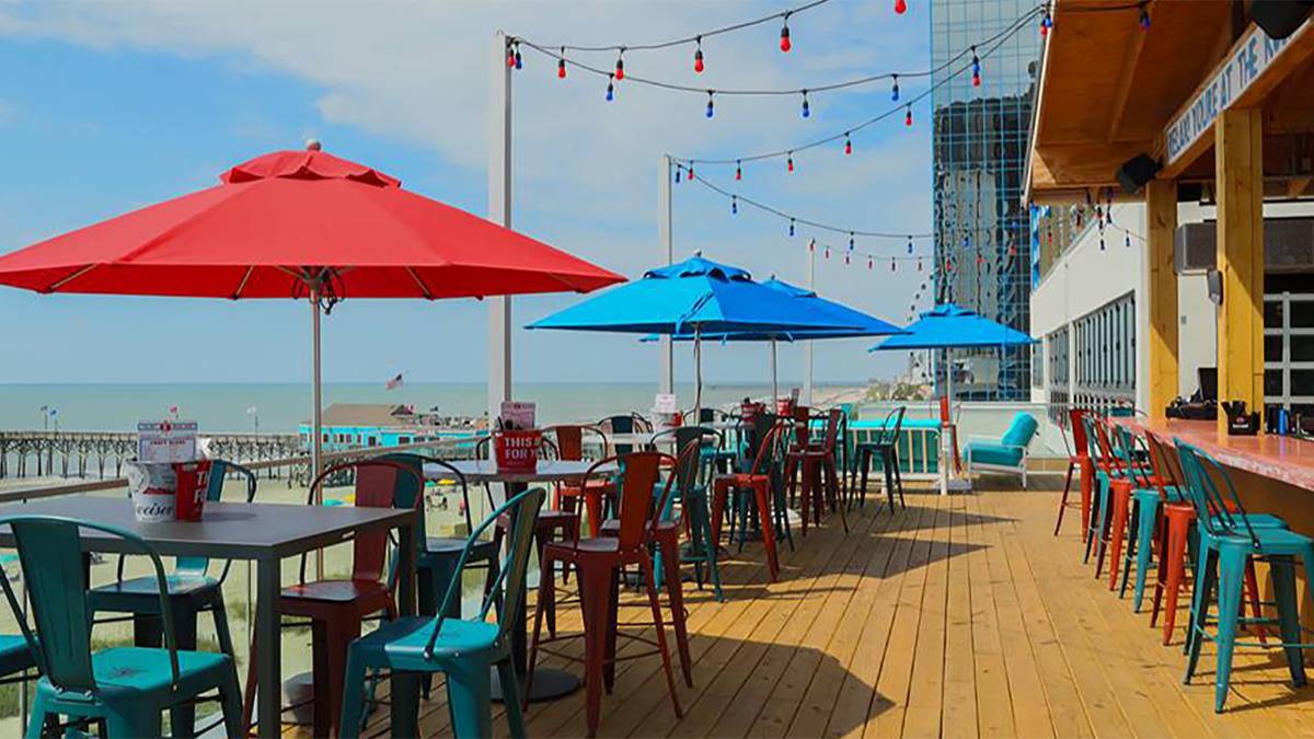 A wooden deck with tall metal tables with blue and red chairs and blue and red umbrella and a bar on the right side at Tin Roof Bar in Myrtle Beach, South Carolina, USA