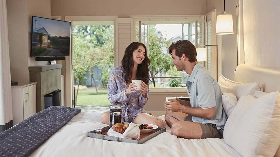 couple sitting on a hotel bed with a breakfast tray and coffee in a bright white decorated room with big open windows behind them