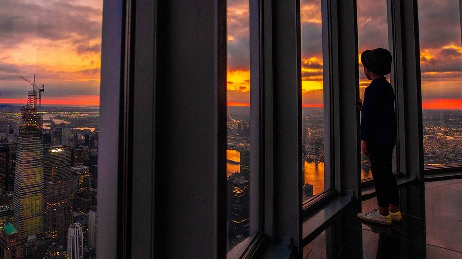 Close up of a person with a hat on looking out the large framed windows of the 102nd Observatory in the Empire State Building at sunset in NCY, New York, USA