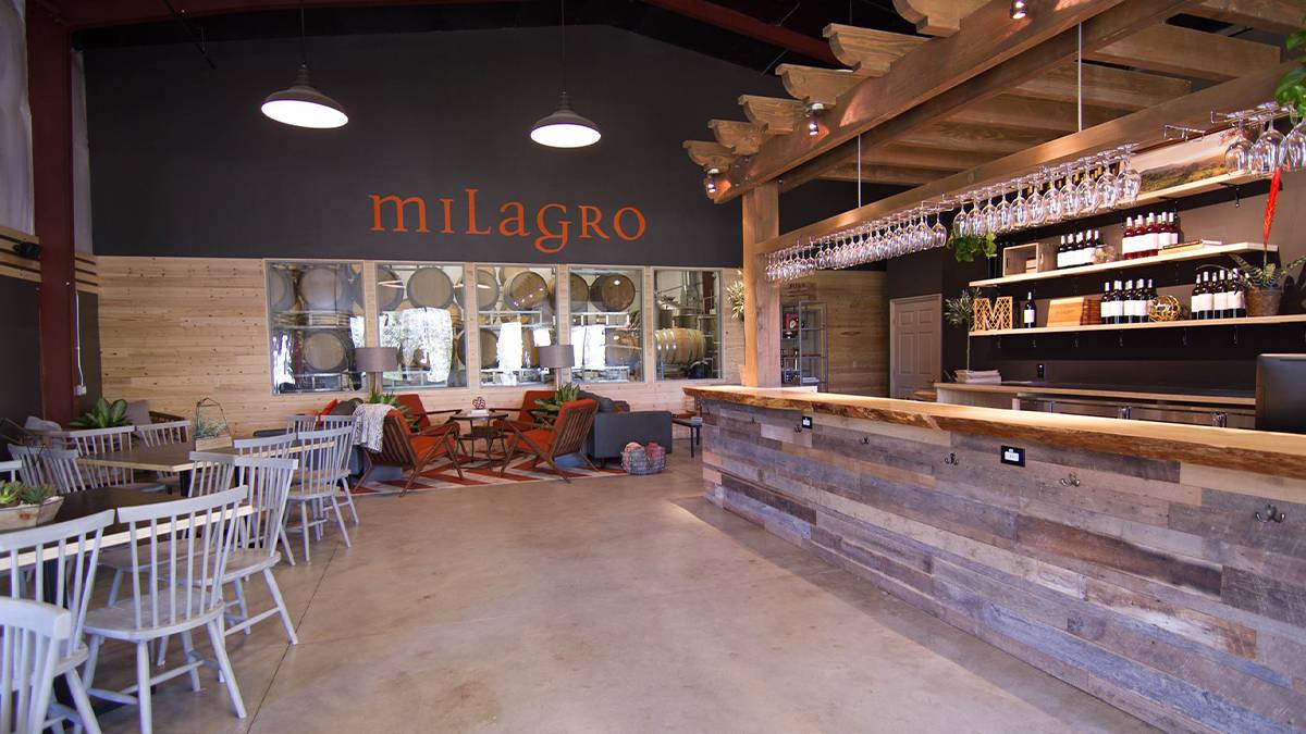 A wooden bar with wine glasses handing from the top and seating on the left hand side with a sign on the back wall in orange saying Milagro at Milagro Winery in San Diego, California, USA