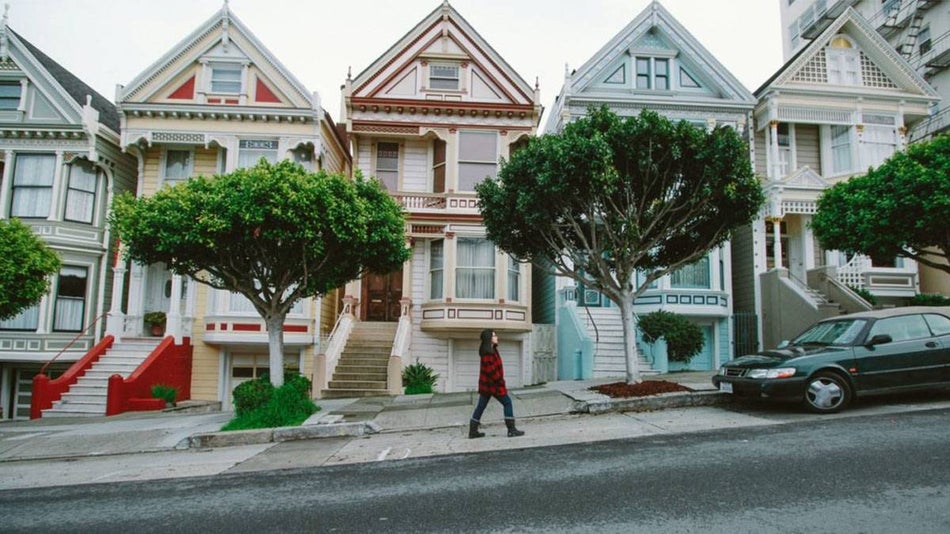 woman in coat and boots walking by the Painted Ladies houses with green trees in front in San Francisco, California, USA