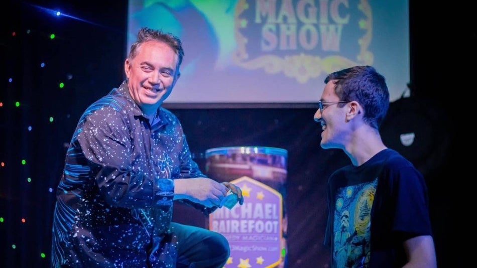Magician on stage performing a trick with a member of the audience sitting next to him