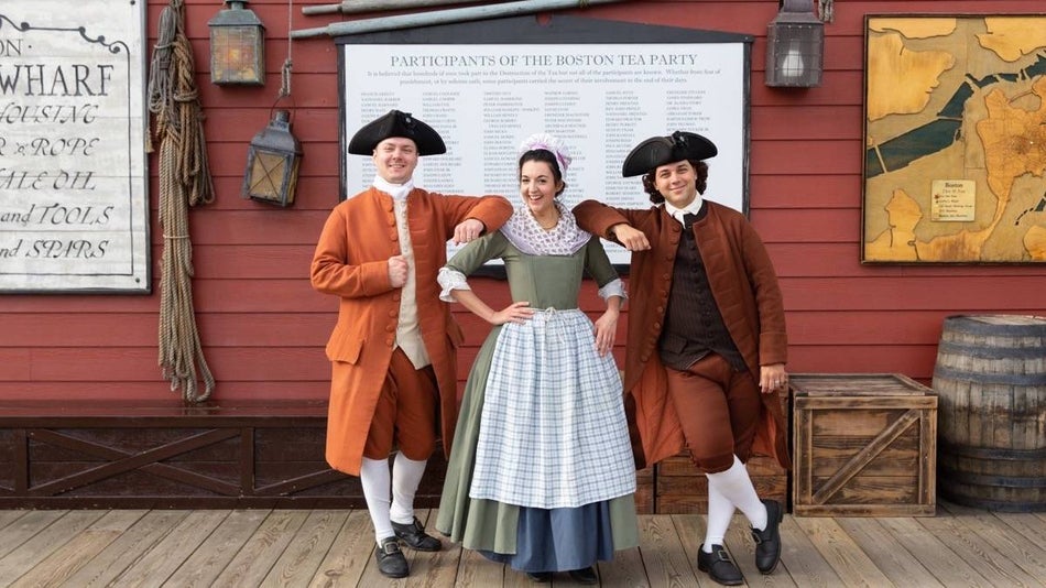 Three people dressed in colonial era outfits standing on a boardwalk with Boston tea party signage behind them
