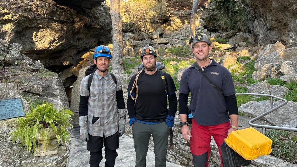 three men with helmets on standing in front of a cave entrance with rocks and some greenery in the background