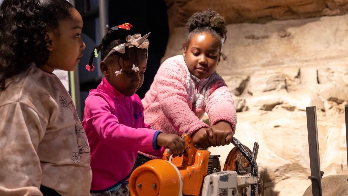 Three small girls in pink tops playing with a mock power tool at an exhibit in the Witte Museum.