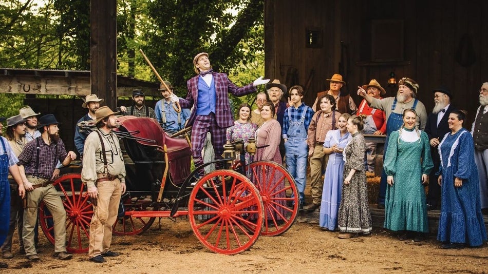Cast members in 1800s costume surrounding a wagon with a man standing on it in a purple suit.