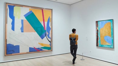 Man in a yellow sweater standing towards the corner of a white-walled room looking at a piece of art hung on the wall at MOMA