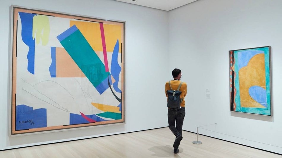Man in a yellow sweater standing towards the corner of a white-walled room looking at a piece of art hung on the wall at MOMA