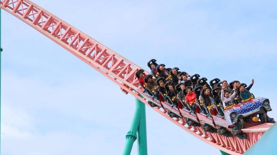Pink track with green supports roller coaster with a car shaped seat with people in it against a blue sky