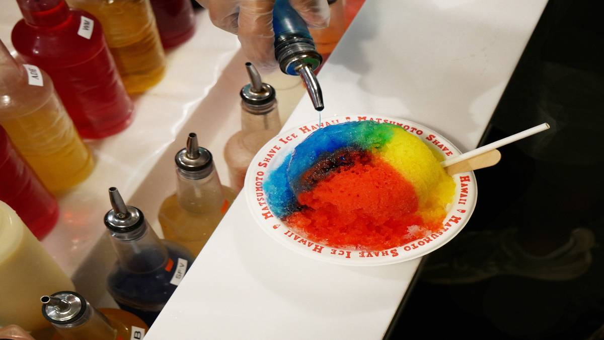 Red, blue, green, and yellow shaved ice on a counter with syrup bottles lined up behind it