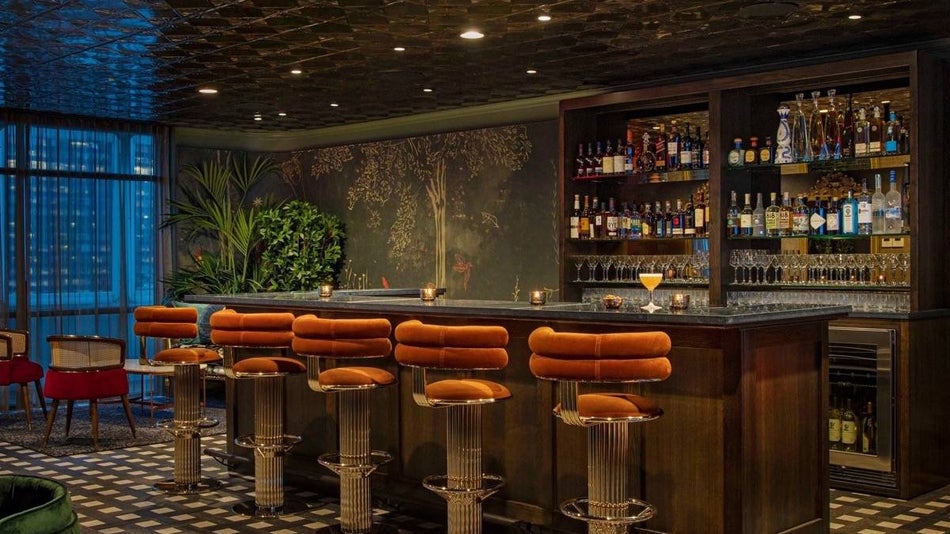 Dimly lit bar area with orange velvet barstools, green floral walpaper and foliage in the corner