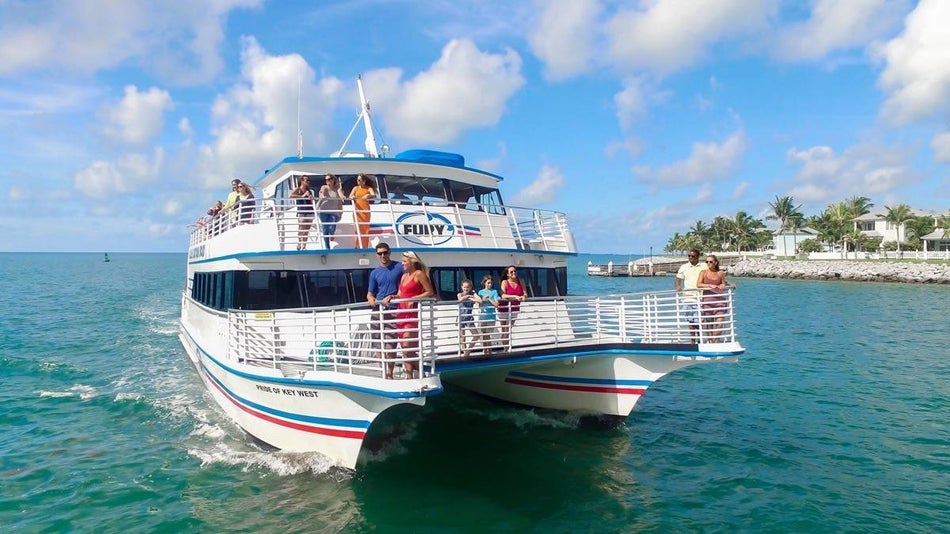 Exterior view of a white catamaran with people sailing with glass bottom boat key west under a blue sky with palm trees on the shore in the background 