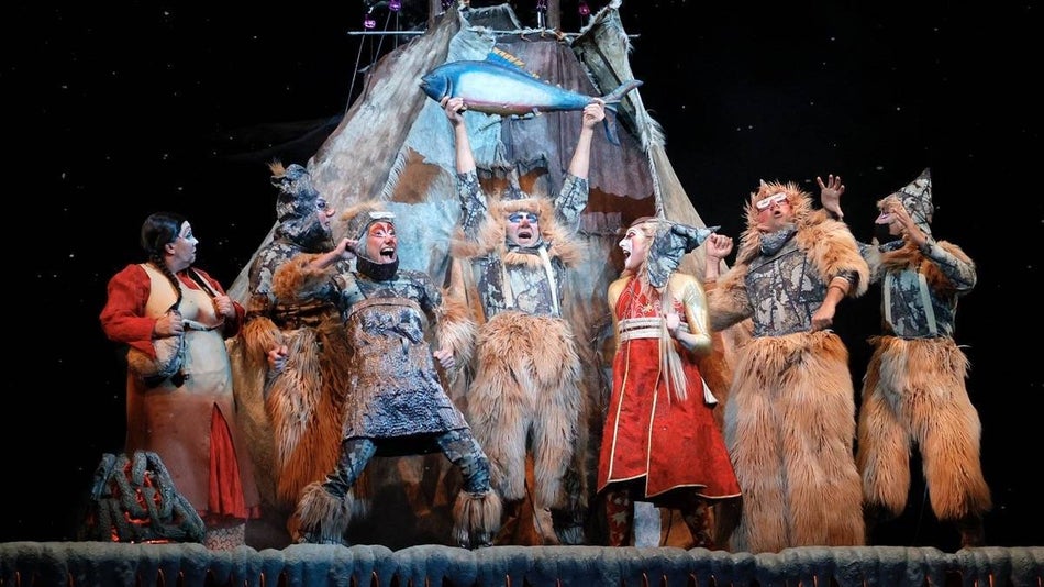 Several cast members of Ka by cirque du soleil standing in furs around a tent while one holds a fish in the air
