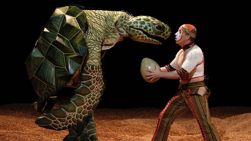 Man holding a turtle egg looking in the face of a giant turtle standing on two legs