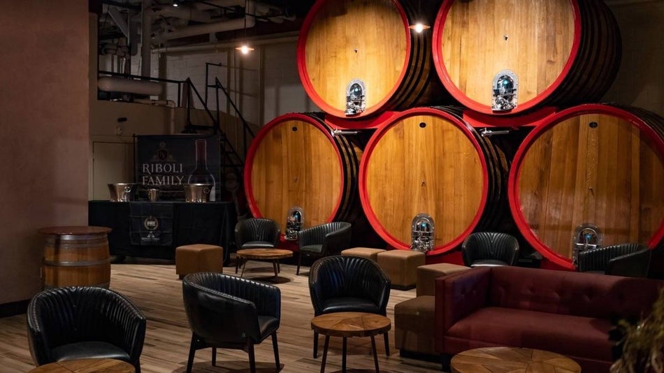 Rows of large wine casks on a wall with tables and chairs in an area for lounging and sipping wine in a dimly lit room