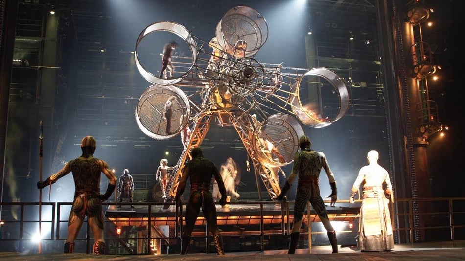 Several cast members of Ka by cirque du soleil watching a spinning large apparatus on stage