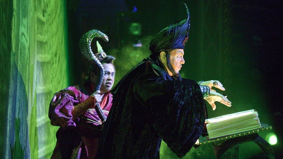 Cast member dressed as Jafar looking menacingly at a book with a green glow cast around him and another cast member looking alarmed behind him