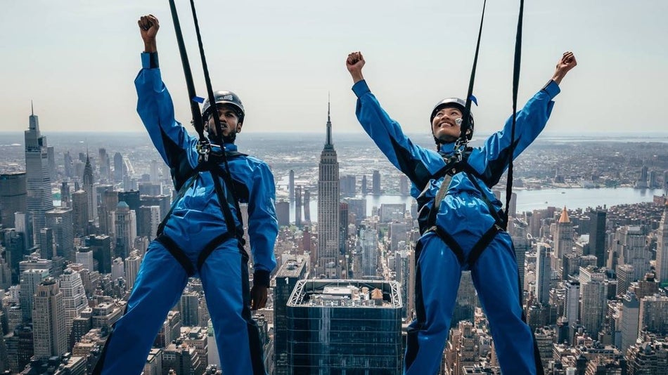 Two people in blue suits in climbing gear with their fists raised leaning back off of a building with the empire state building in the background