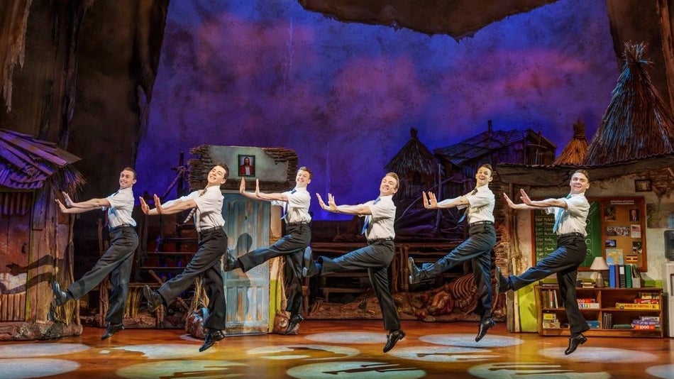 Several cast members in white button down shirts with black pants jumping in a row on stage