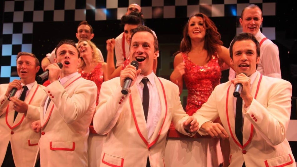 Four men on stage singing with several backup singers behind them