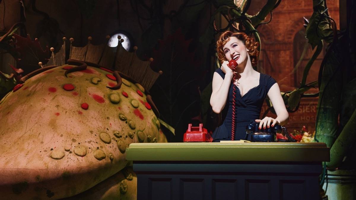 Woman with red hair in a black dress talking on a red phone with a huge plant creature behind her