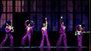 Several cast members on stage in purple suits singing and playing guitar for MJ the musical