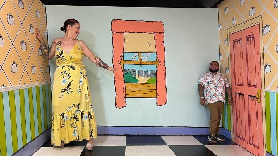 Woman in a yellow dress looking over at a man in a fun house room where they look disproportionately sized