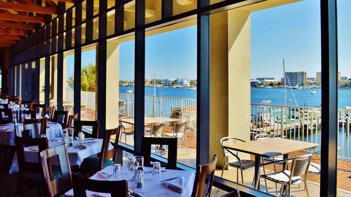 Tables with white table clothes next to large windows overlooking a bay with outdoor seating