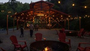 Open barn pavilion with a fire pit, chairs, string lights and people walking around at night