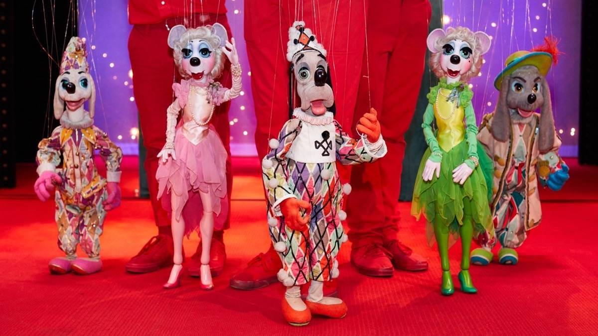 5 brightly costumed dog puppets with three people holding them up in red pants and red shoes