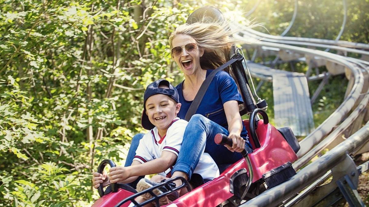 Woman and little boy riding on a red coaster car on a metal track with greenery all around them