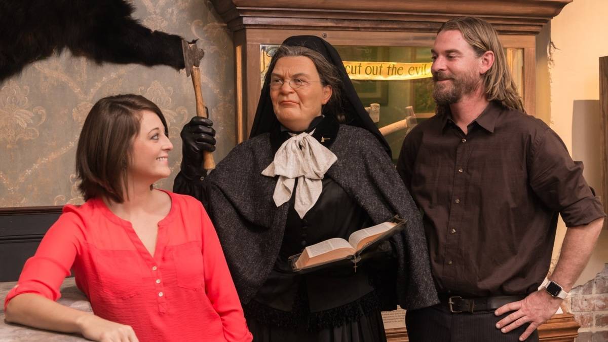 Wax figure woman in black holding a book and an ax with two other visitors in the photo