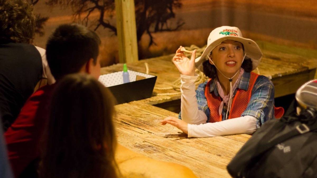 Woman in a bucket hat holding a small bone at a wooden table while children watch her