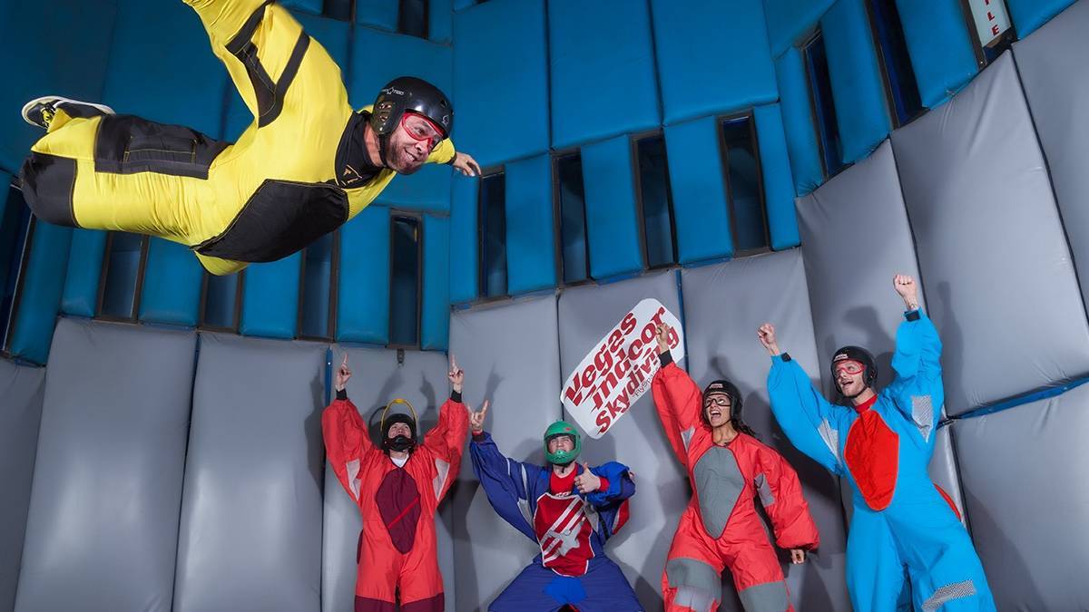 one person in a flight suit skydiving indoors with four people behind them cheering them on