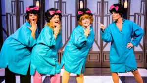 Four women in blue salon capes with black wigs on and pink bows in their hari