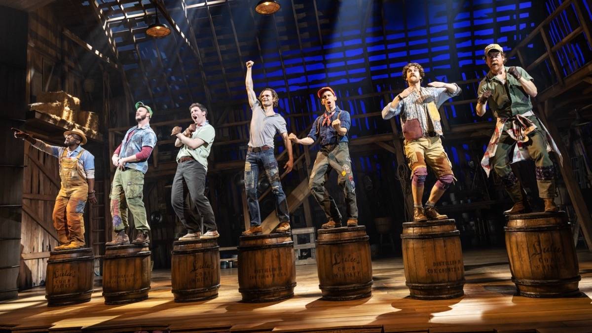 Seven men in farm attire dancing on top of wooden barrels in a barn