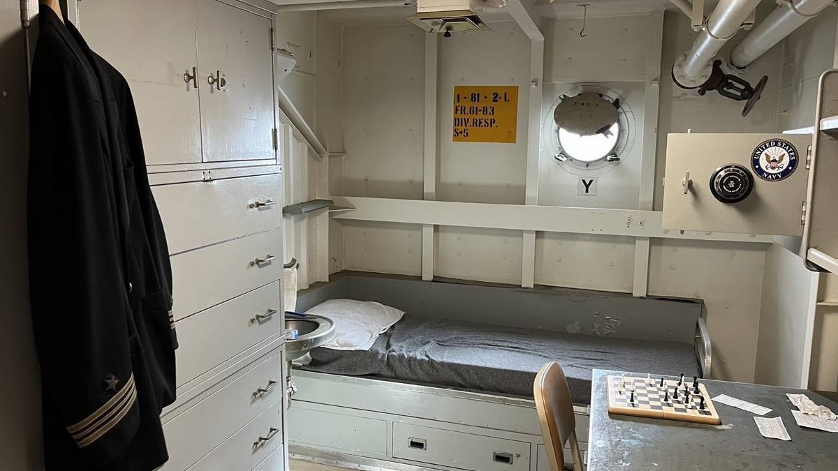 Interior cabin of a battleship with a small cot, white washed walls, a desk with a chess game and a uniform hanging from a closet door