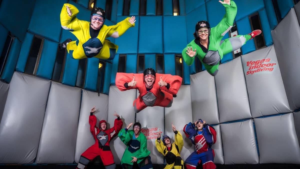 Three people in flight suits skydiving indoors with four people behind them cheering them on