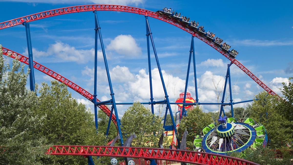 Red roller coaster with blue supports over green trees and a blue and green spinning ride
