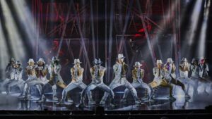 14 performers in white suits singing on stage