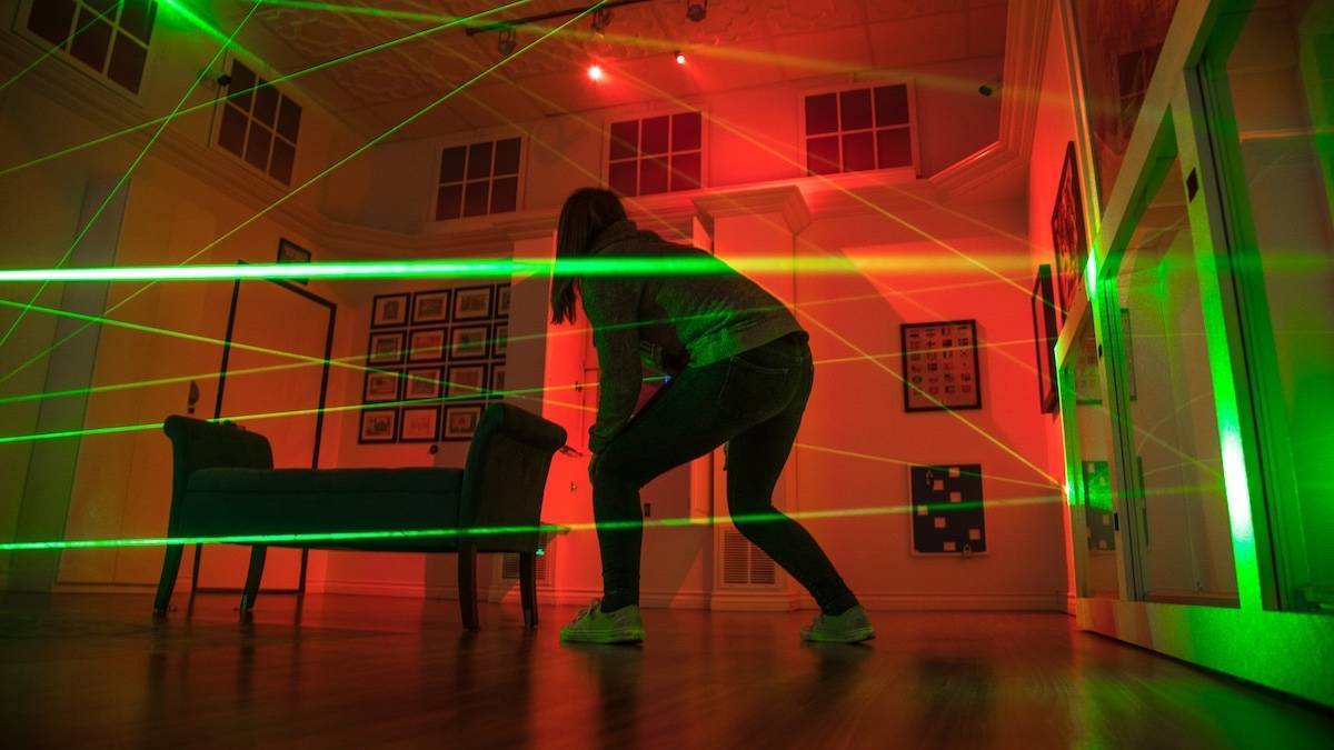 Girl crouching in a red lit room criss crossed with green laser beams