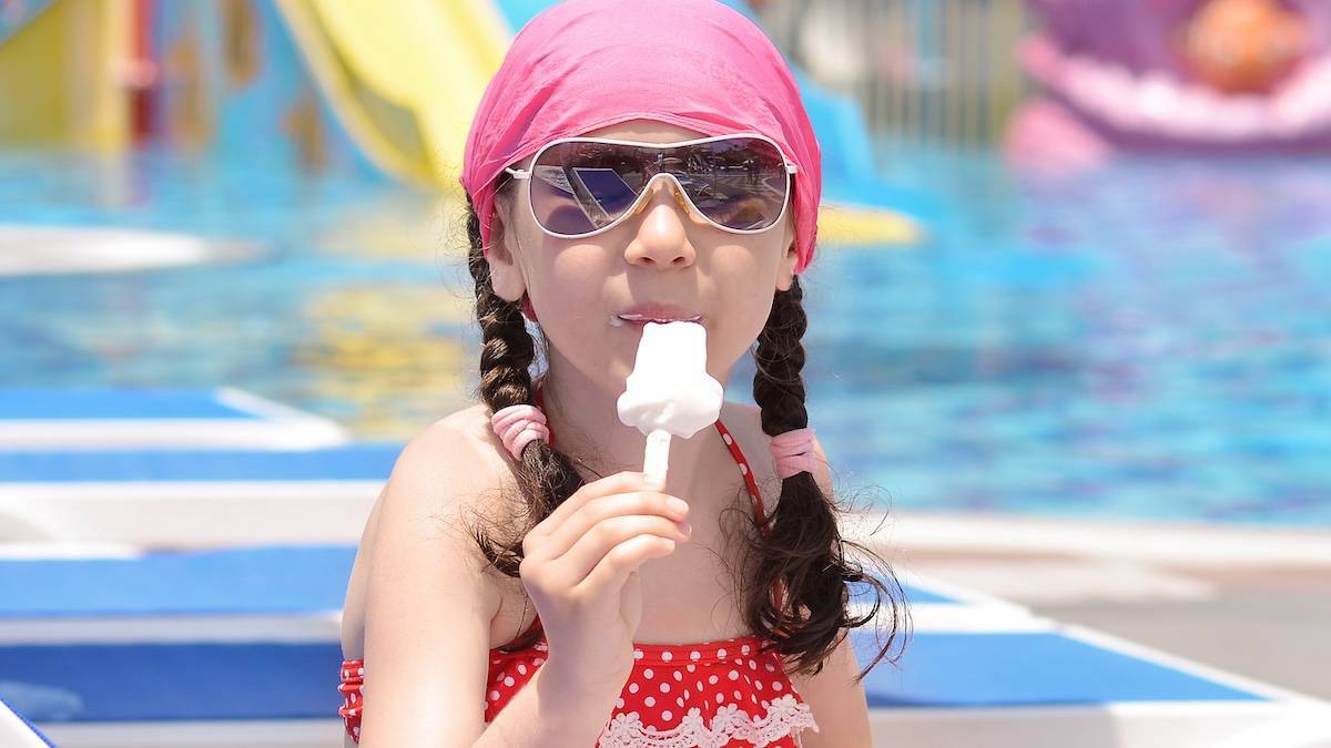 Girl with brown hair in braids with a pink bandana on eating an ice cream with a red polka dot swim suit on