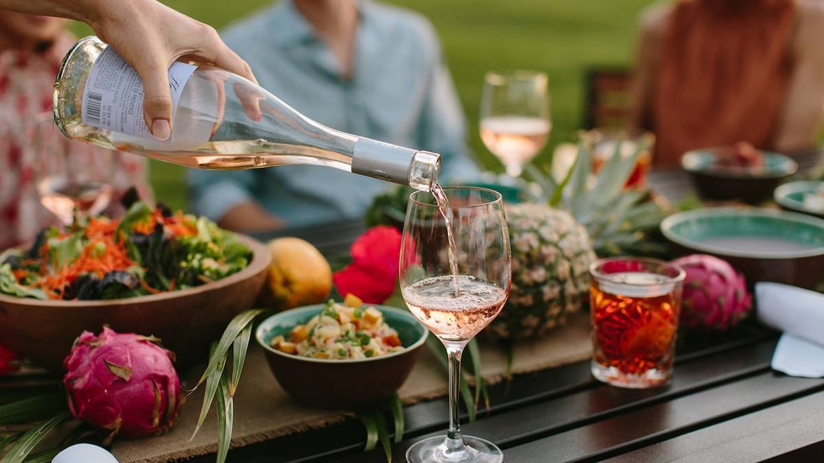 Wooden table laden with fresh fruits and salad and wine glasses as someone pours rose wine into a glass