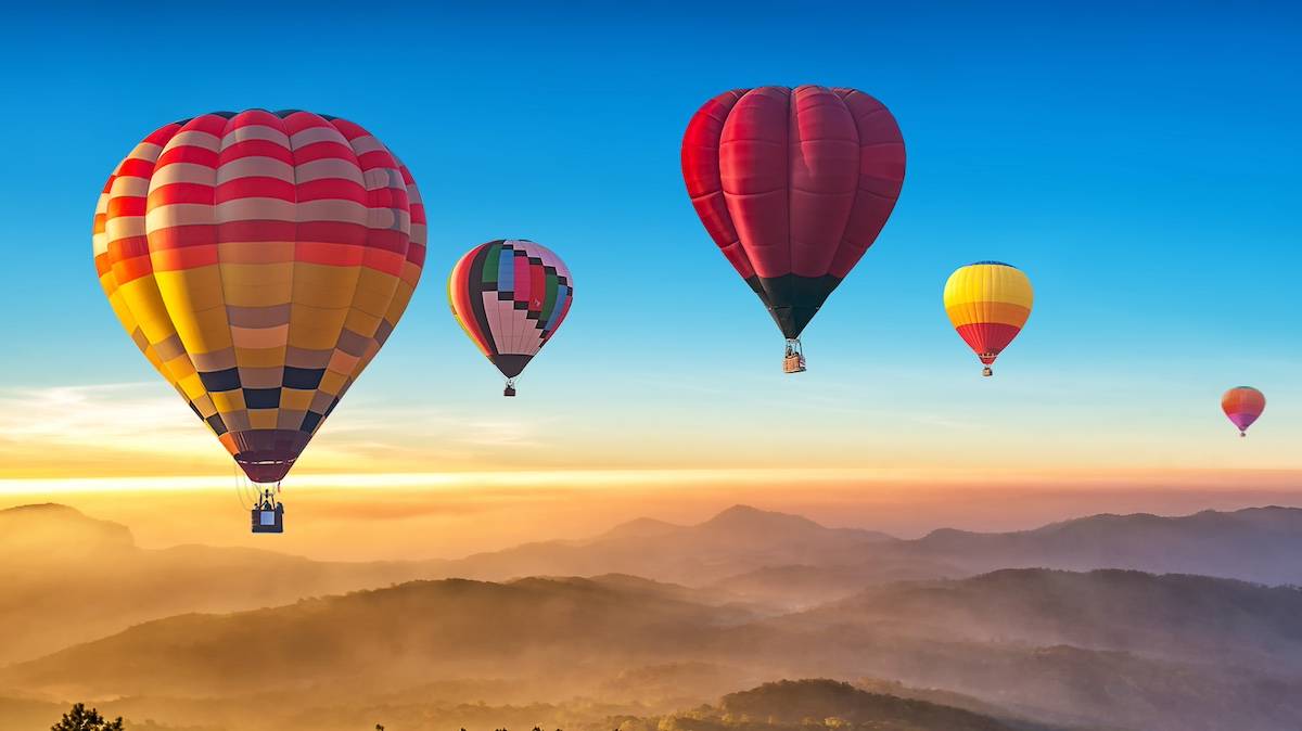 Several colorful hot air balloons in the sky over a cloudy mountain range at sunrise