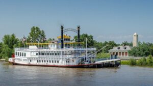 Large cruise boat named Creole Queen on the water with a plantation house and a bright blue sky behind it in New Orleans, Louisiana