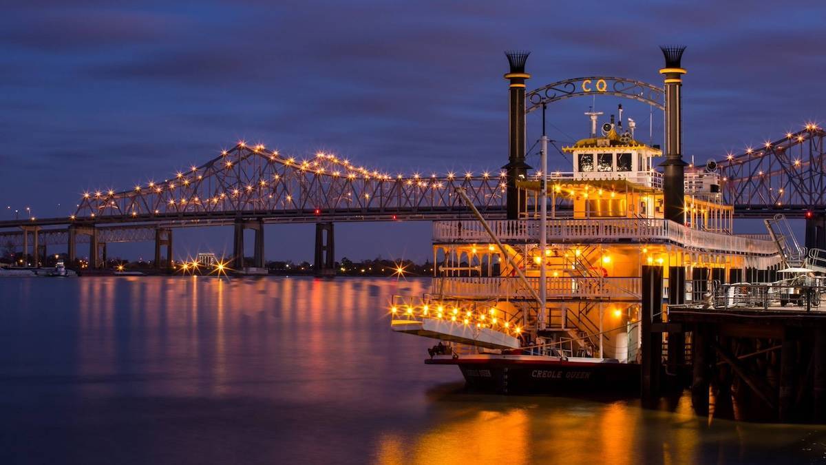 Large cruise boat named Creole Queen on the water with a bridge and a cloudy evening sky behind it in New Orleans, Louisiana