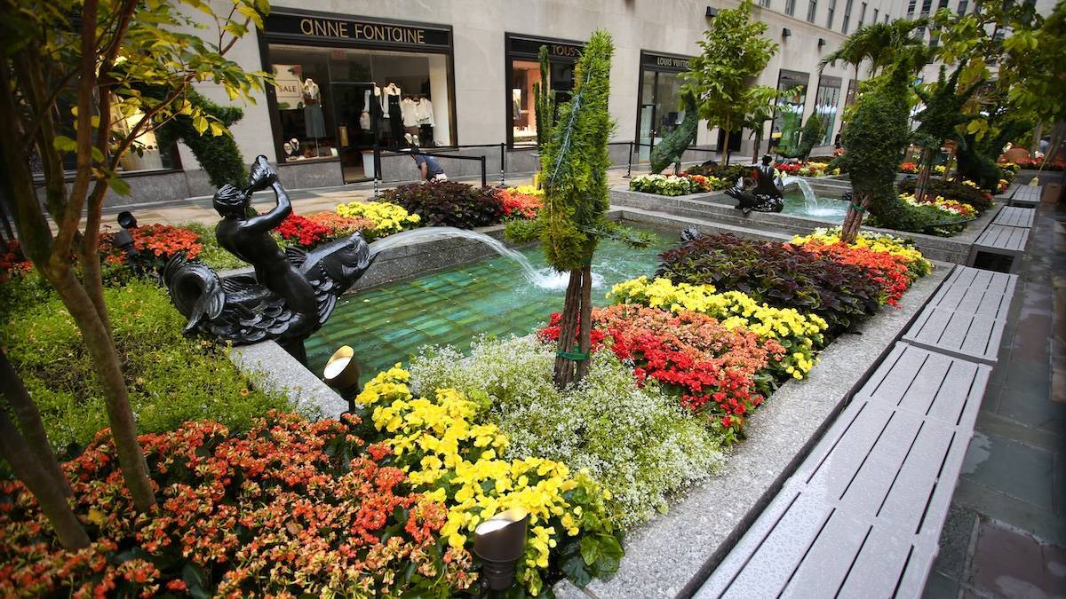 Colorful flowers and greenery with a small fountain and statue surrounded by benches at Rockefeller Center in NYC