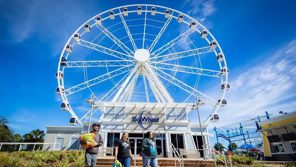 Close shot looking up at a white ferris wheel under a blue sky with three people walking in front of it